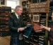 A man wearing a tweed jacket and kilt examining fabric swatches with a shopkeeper inside a traditional Scottish tailor shop filled with bolts of tartan.